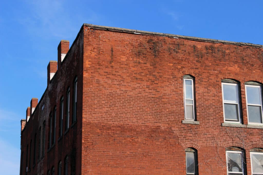 A corner view of a red brick building showing the side facade with several windows. The structure has a slightly weathered appearance, and the blue sky is visible above with a few scattered clouds.