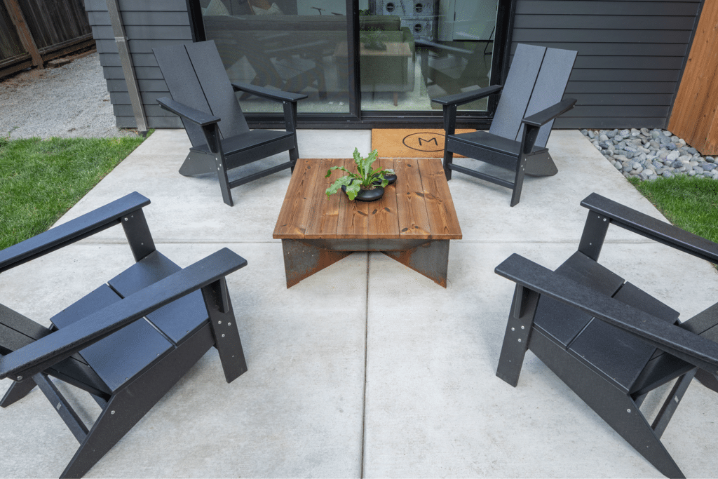 AdobeStock_475771681 1 A small outdoor seating area featuring four black Adirondack chairs arranged around a wooden coffee table with a plant centerpiece. The concrete surface is surrounded by grass and a wooden wall in the background.