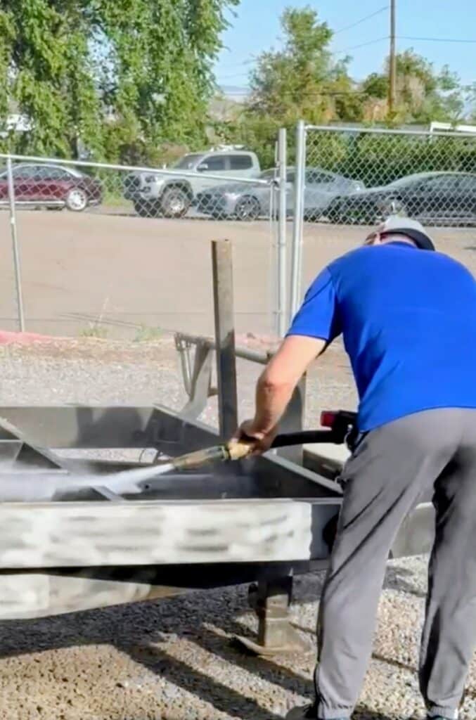 Screenshot_20260713_084559_Messages~2 A man in a blue shirt and gray pants washes a large metal grill outdoors beside a chain-link fence, with parked cars and trees in the background.