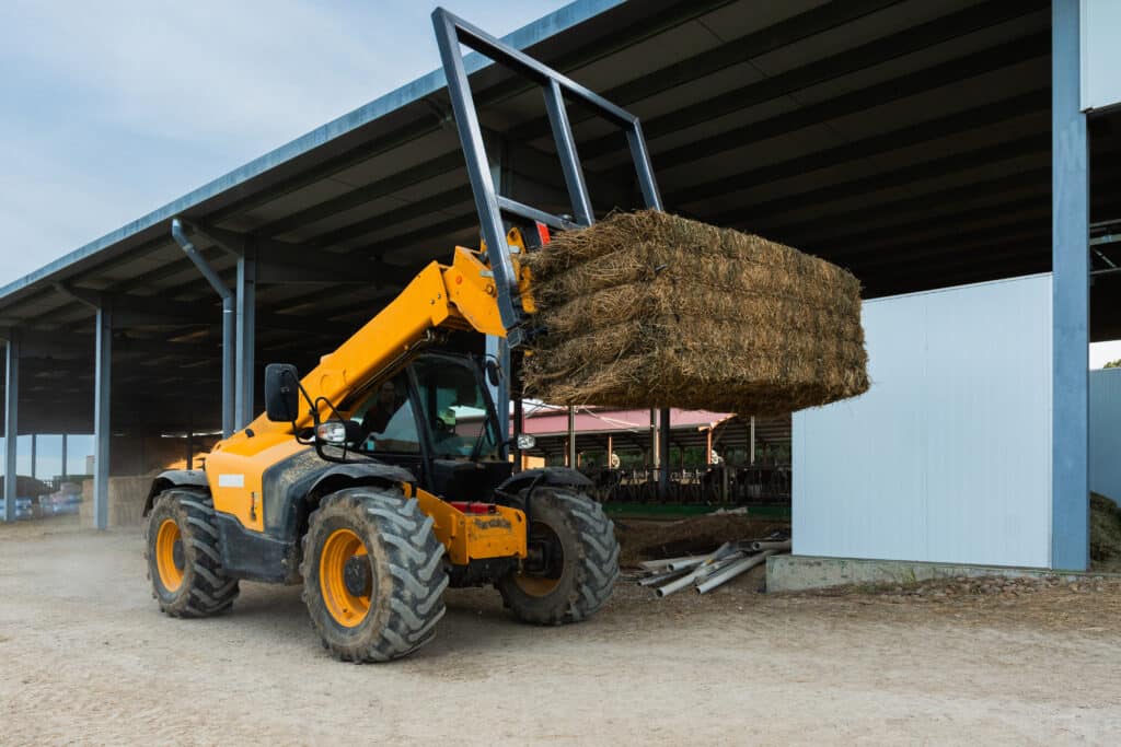 Telehandler lifting hay bale in agricultural farm building A yellow telehandler lifts a large stack of hay bales with raised forks beside a metal agricultural shed.